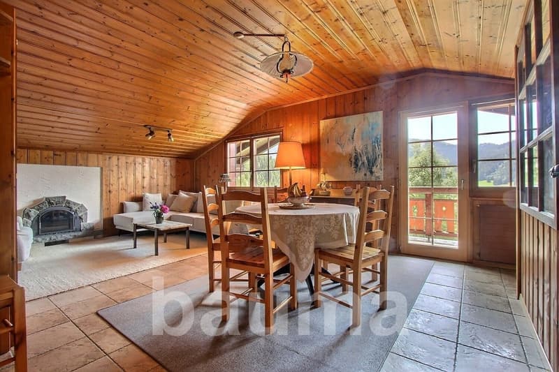 Spacious open-plan living and dining area with wooden paneled walls and ceiling, large windows offering a scenic view, a fireplace, a dining table with chairs, and a sofa set in the living room area.