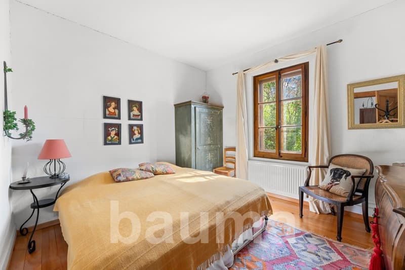 A spacious bedroom with a bed, a bedside table with a lamp, a chair, a cabinet, a radiator, and a mirror above the bed.