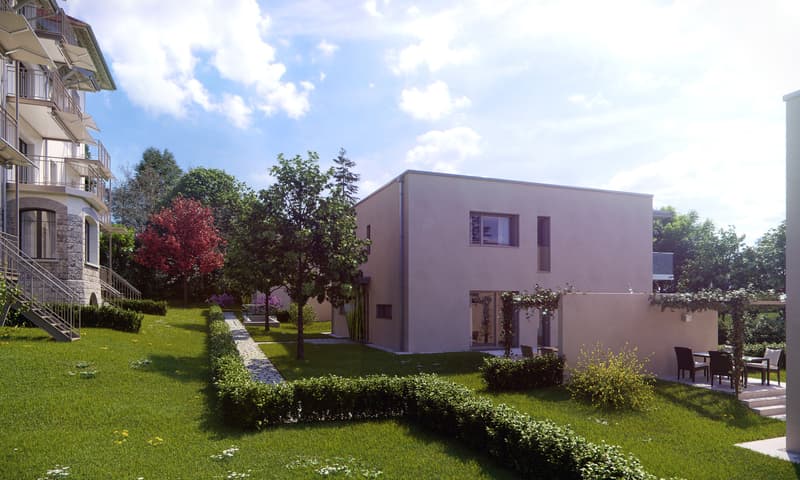 Modern two-story residential building with balconies, surrounded by a well-maintained garden with trees, shrubs, and a paved walkway. The building has a simple, minimalist design with a neutral color scheme and large windows. There appears to be a parking