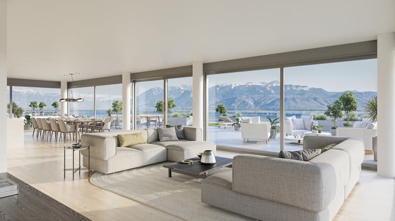 Spacious open-plan living area with large windows offering panoramic views of the mountains and lake. The room features a comfortable seating area with plush sofas and armchairs, a dining table, and a modern kitchen visible in the background. The decor is