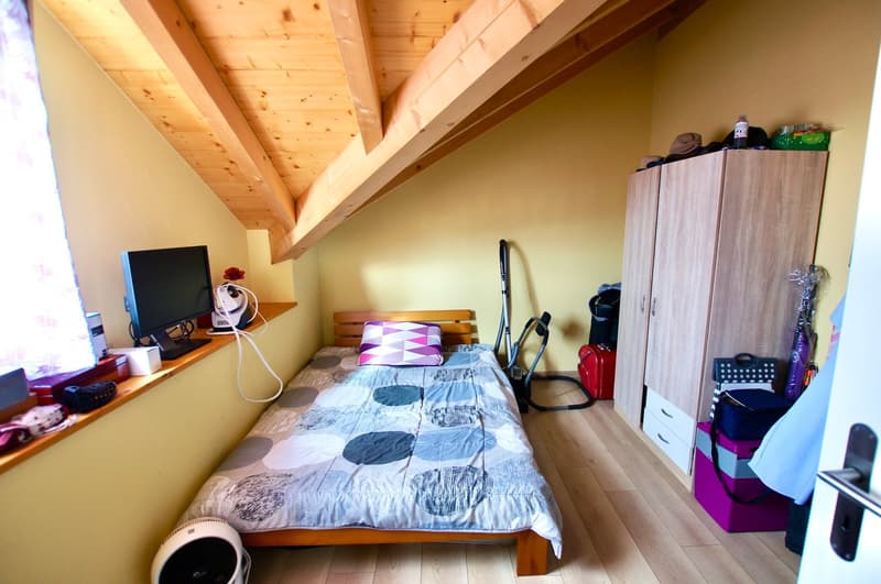 Small bedroom with bed, desk, window, and wardrobe. The bed has a pillow and blanket. A desk holds a monitor, speaker, and storage box. The window includes curtains and a flower. The wardrobe is filled with bags and hats.