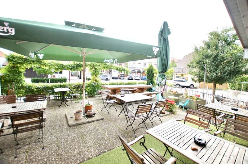 Outdoor seating area with tables and chairs, green umbrellas, plants, and a view of the street.