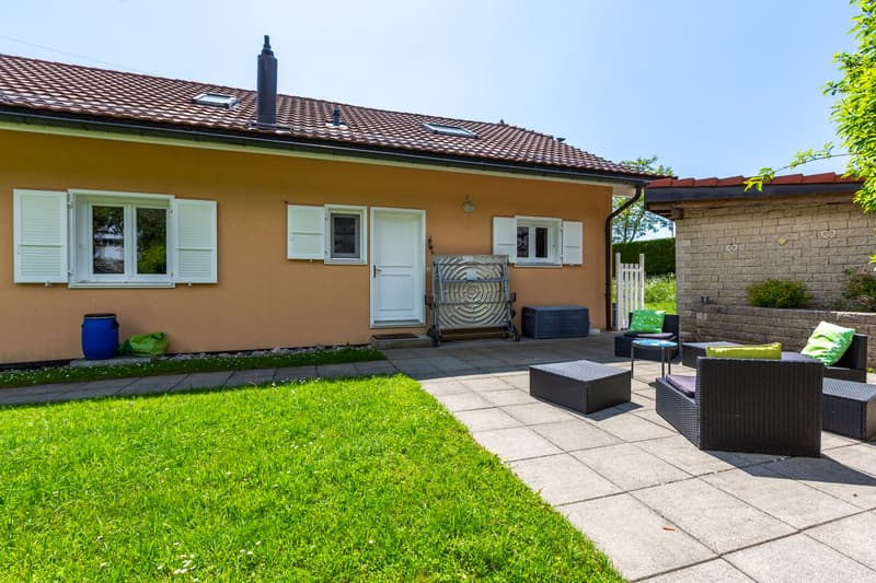 Single story house, brown roof, white shutters, tiled outdoor seating area, storage box, potted plants, barrel, grass lawn, patio table, patio chairs