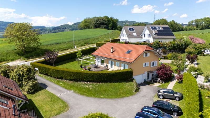 House with red roof, surrounded by bushes, balcony, several cars parked, detached garage, green field background.