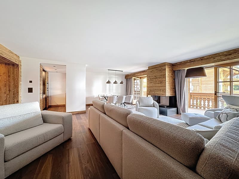Modern living room with wooden flooring, white walls, glass windows with balcony, chandeliers, sofas, TV, dining table, chairs.
