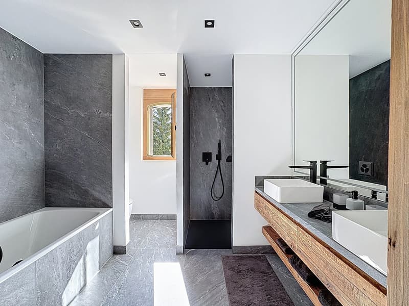 The bathroom has a bathtub, two sinks, a mirror, a wooden countertop, and a small window leading to the outdoors.