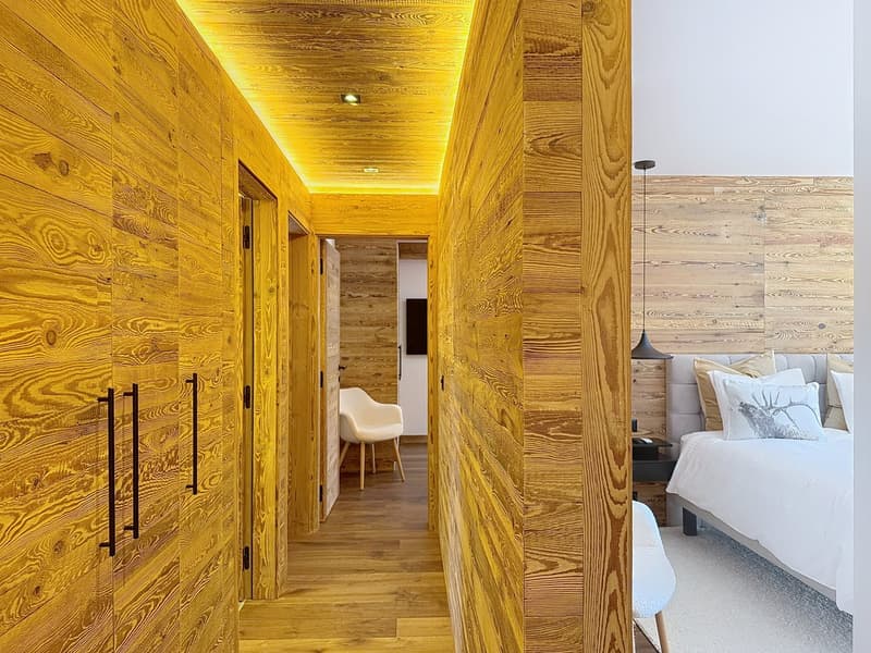 Wooden walls, illuminated hallway, modern chair, bedside table, bed with white sheets and multiple decorative pillows