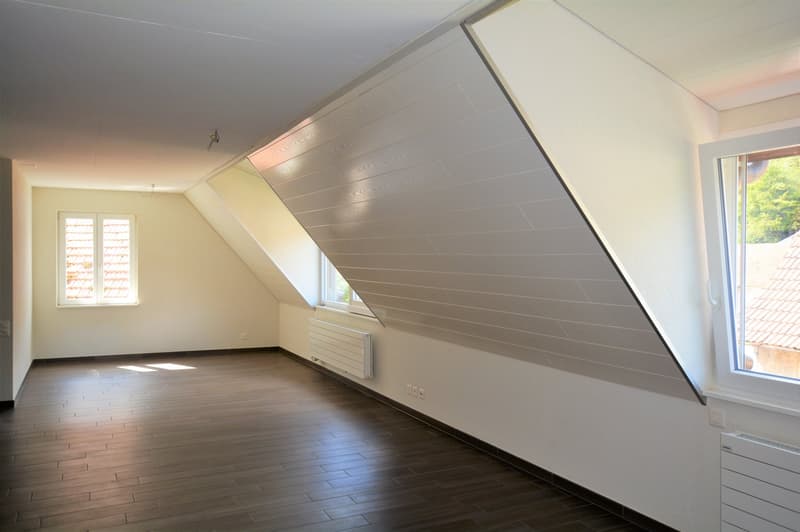 Spacious attic room with slanted walls, hardwood flooring, and large window providing natural light. The room appears to be unfinished, with exposed ceiling beams and minimal furnishings.