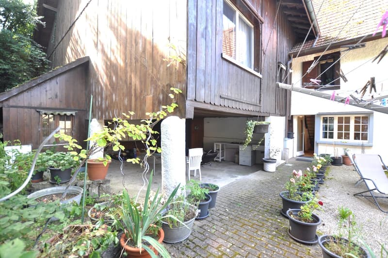 Two-story wooden house with a porch, surrounded by potted plants and greenery. The house has a rustic, rural appearance with a brick paved pathway leading to the entrance.