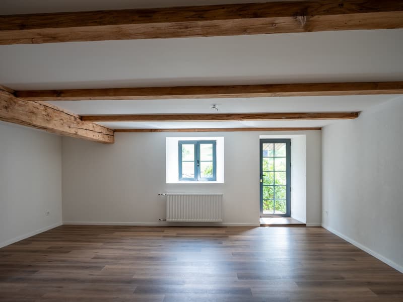Empty room with wooden floors, white walls, wooden beams, window, door, radiator.