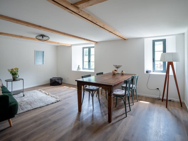 Dining room with wooden floor, dining table with chairs, green couch, wooden ceiling beams, floor lamp, rug, side table with vase, picture on wall