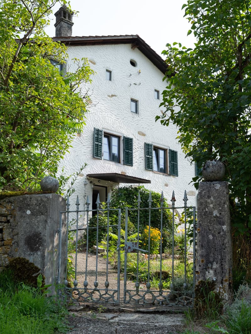 A large white building with stone walls, a large entrance gate, two stories, a chimney, and green shutters.