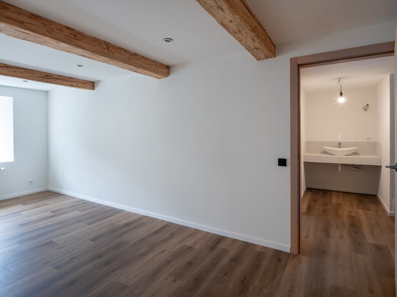 Empty room with wooden flooring, white walls, wooden beams, power outlet, bathroom sink, ceiling lamp