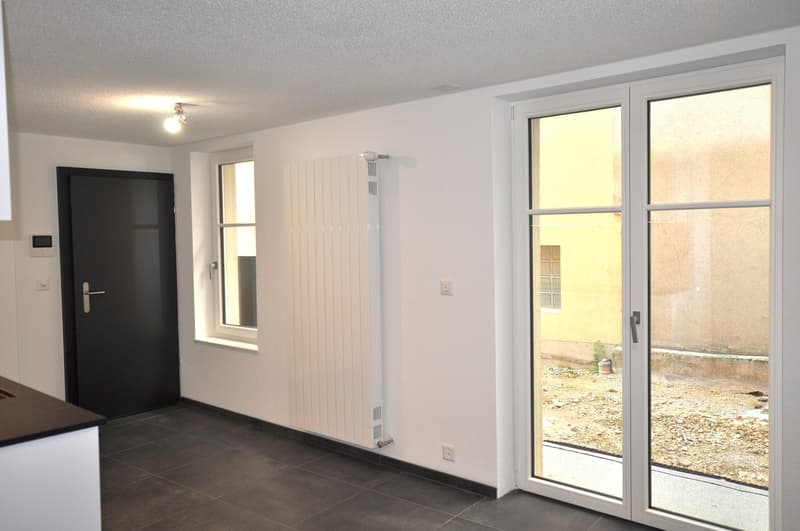 An empty room with a white radiator, tiled floor, white walls, sliding doors to a terrace, small window with wall-mounted heater, entrance door.