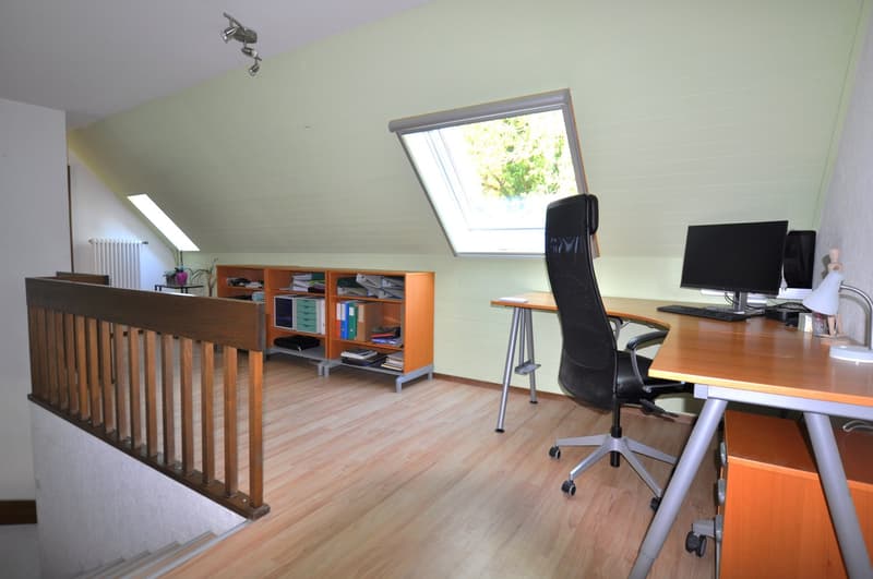Office with desk, monitor, keyboard, lamp, computer chair, shelves with books and office supplies, attic window, radiator, wooden floor