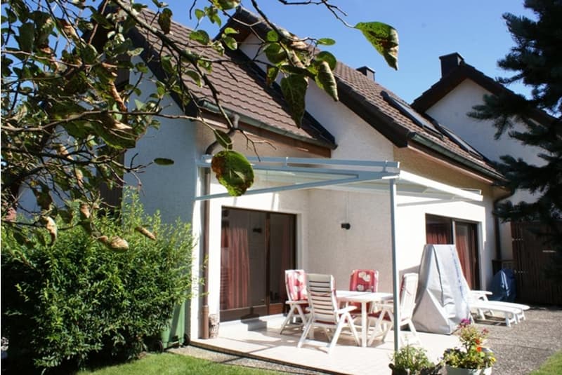 Detached house with brown roof, white walls, covered patio with table and chairs, outdoor cover, and a garden with plants.