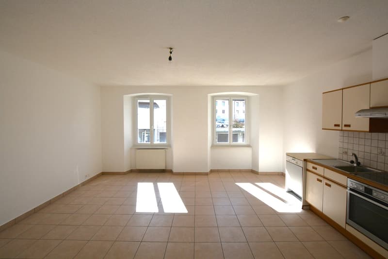 Open room, two windows, light fixtures, light wooden cupboards, white tiles on floor, kitchen appliances