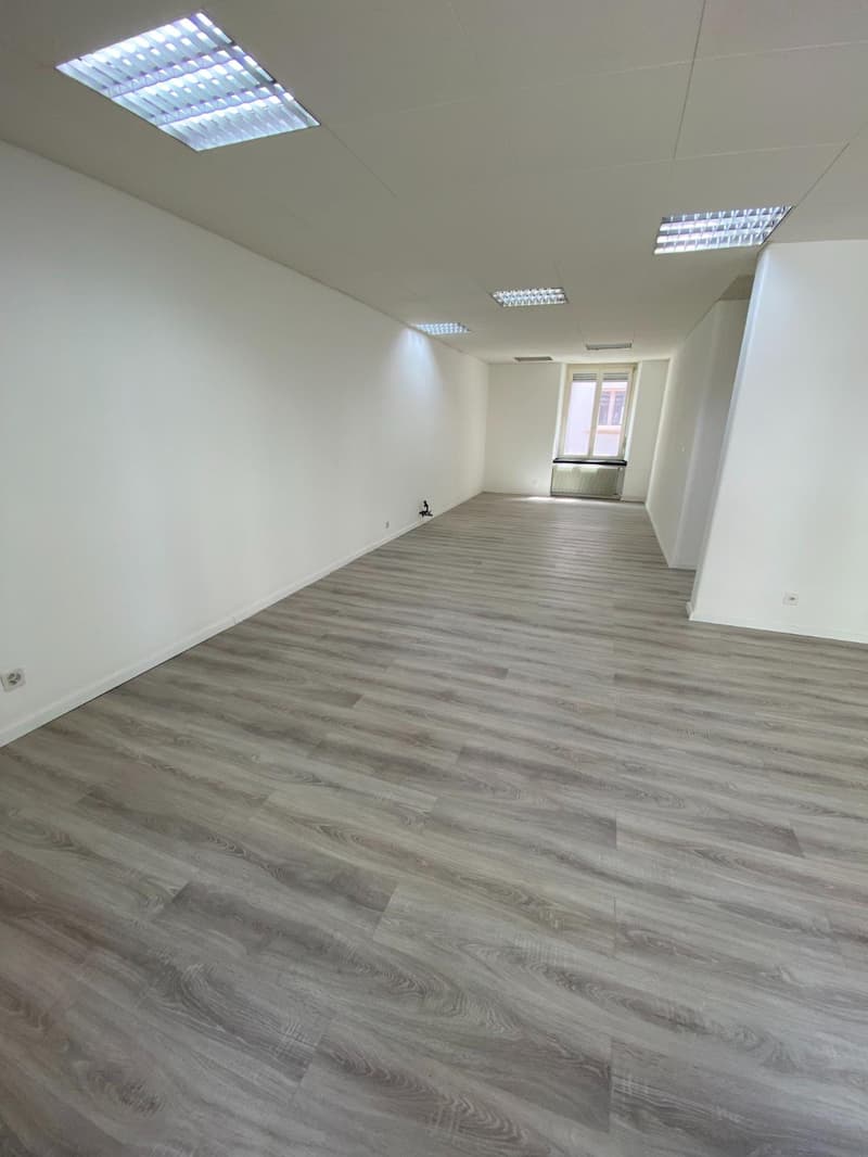 An empty room with wooden flooring, white walls, fluorescent lights, and an open window with an exterior view. The room appears to be a blank canvas, ready for customization or use. The wooden flooring adds a touch of warmth and elegance to the space, whi