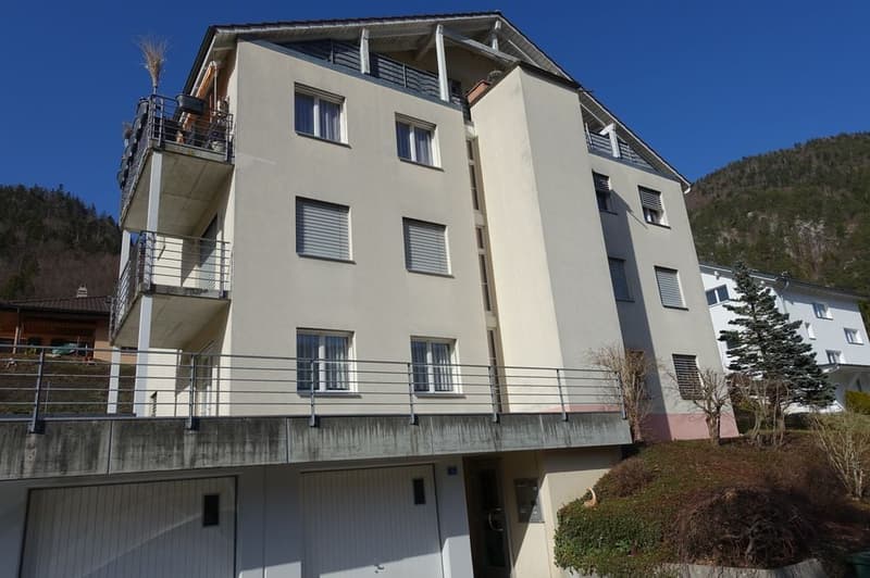 Apartment building with multiple floors, balconies on each floor, garage doors, and a small courtyard area in front.
