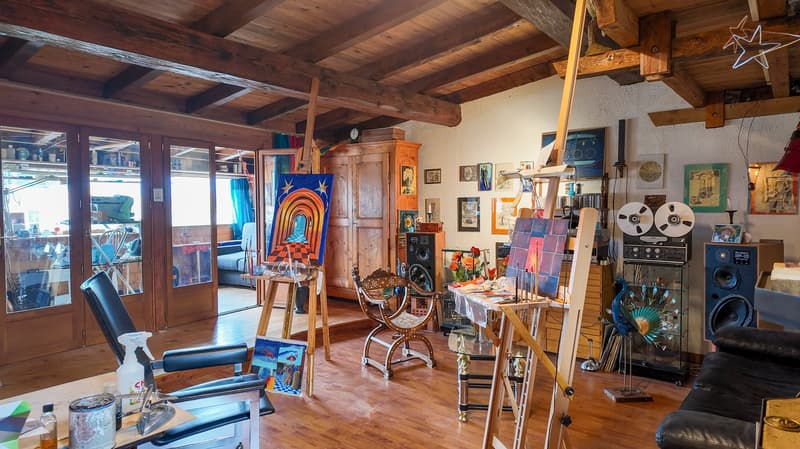 wooden floor, wooden beam, easel with artwork, paintings on the wall, speakers, armchair, black leather couch, table with painting supplies