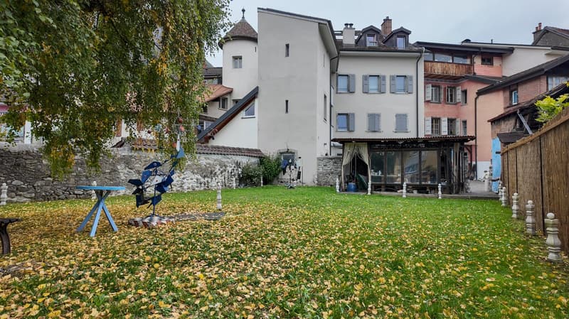 A grassy garden area with fallen leaves, a stone wall, a blue table with a small sculpture, a modern sculpture, and several buildings in the background.