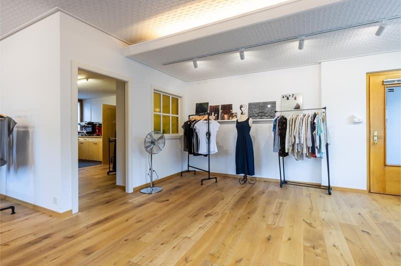 a room with white walls, wooden floor, clothes hanging on racks, clothes hanging on a wall, a fan, a cabinet, and pictures on the wall