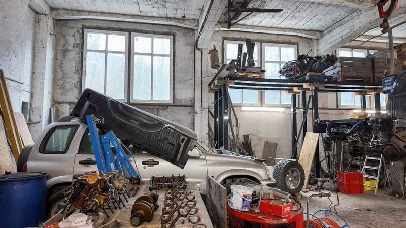 A workshop space with a car being serviced, multiple engine parts and tools scattered on the ground, and a lift in the background.