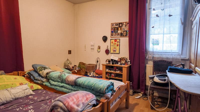 two beds with multiple pillows, books, a table with a heater, a window with white curtain, and a small cabinet