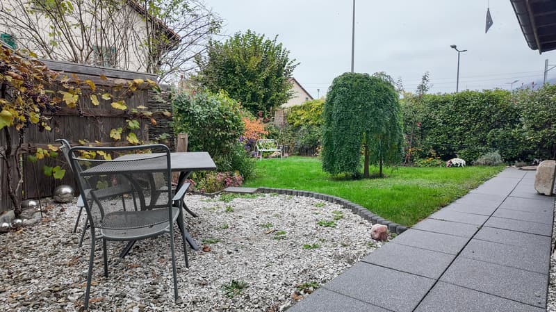 gravel area with table and chairs, green lawn with plants and flowers, a small white chair in the garden, a wooden fence with a grapevine on it, a pathway leading to the house, a small silver object