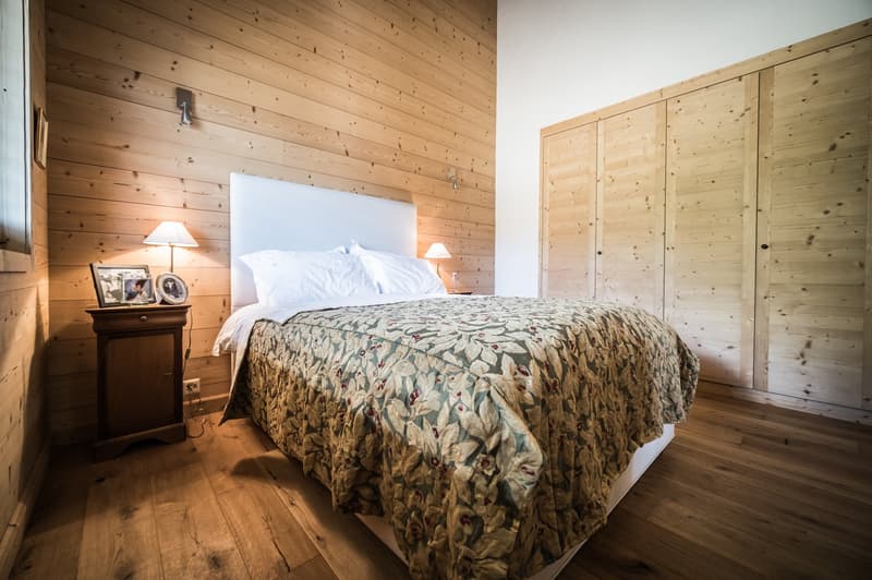 wooden bed, wooden nightstand with two lamps, three white pillows, wooden bedspread, two closets with wooden doors, picture frames, white wall