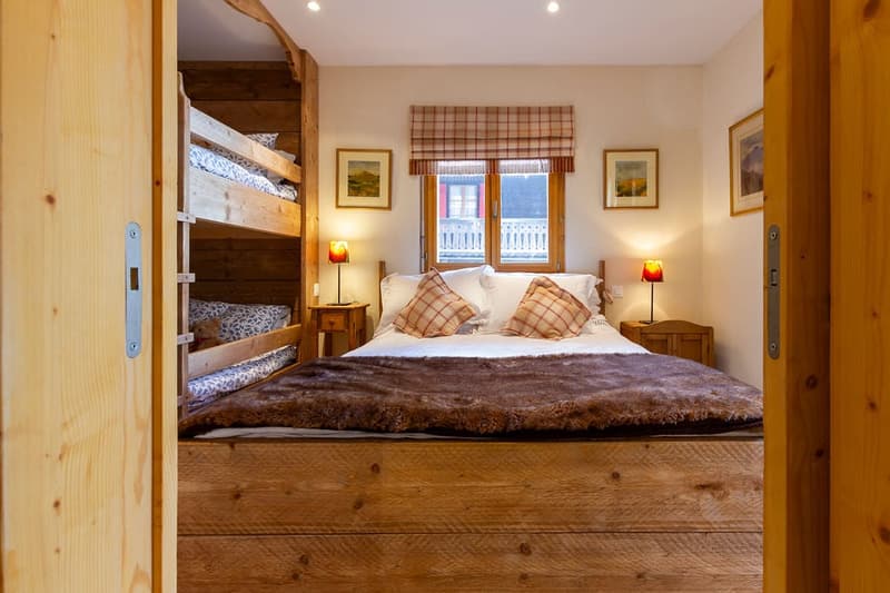 Cozy bedroom with wooden bunk beds, large bed with fur blanket, wooden furniture, and plaid curtains overlooking a balcony or terrace