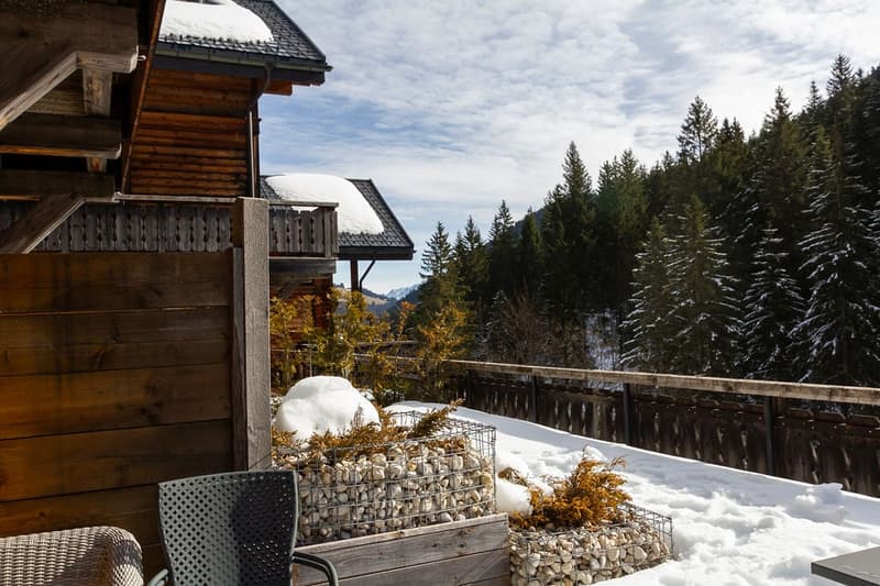 Wooden cabin with a balcony/terrace, surrounded by snow-covered pine trees and mountains in the background. The cabin has a rustic, cozy appearance with a stone wall feature on the balcony.