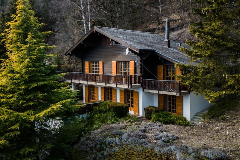 A rustic wooden cabin or chalet nestled in a forested mountain setting, with a large balcony or terrace overlooking the surrounding landscape. The cabin has a slate roof and wooden shutters on the windows, blending in with the natural environment.
