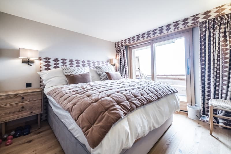 Double bed with wooden headboard, several pillows, white walls, beige wooden floor, night lamps, bedside cabinet with drawers, open glass door with balcony access, small trash bin, decorative stool, small exercise ball