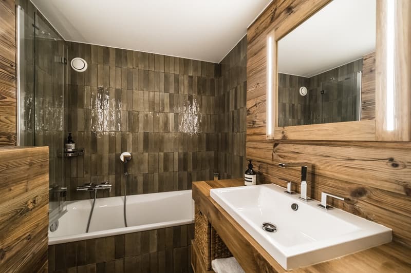 Wooden walls, white bathtub, glass shower, wooden vanity with sink, mirror, wicker baskets, shower faucets, soap dispenser