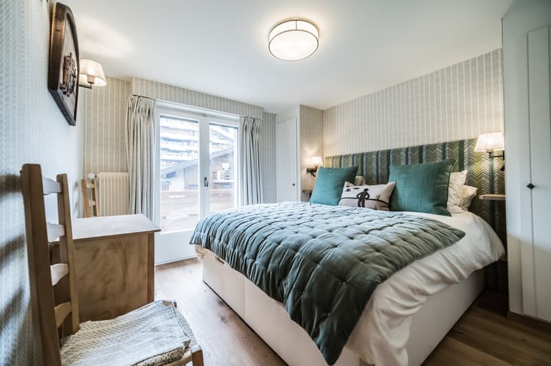Wooden flooring, bed with green blanket, pillows, radiator on the wall, wooden chair, ceiling lamp, wall lamps, window with curtains, wall mounted frame