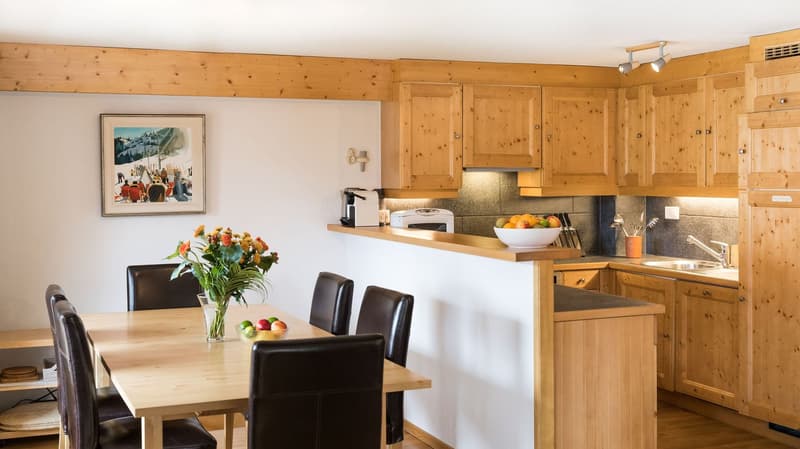 Modern design kitchen, wooden cabinets, stainless steel appliances, countertop with bar stool seating, sink with faucet, refrigerator, coffee machine, toaster, dining table with chairs, picture frame, flowers, wooden flooring.