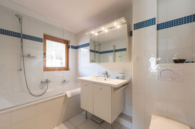 Bathroom with white tiles, glass shower enclosure, bathtub, white vanity, sink, mirror, and window.