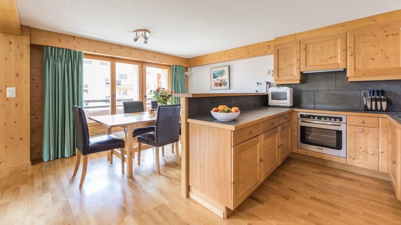 Open kitchen, dining table with chairs, wooden cabinets, white countertop, stainless steel oven, microwave, bowl with fruits, green curtains, picture frame, electrical outlet, wooden flooring