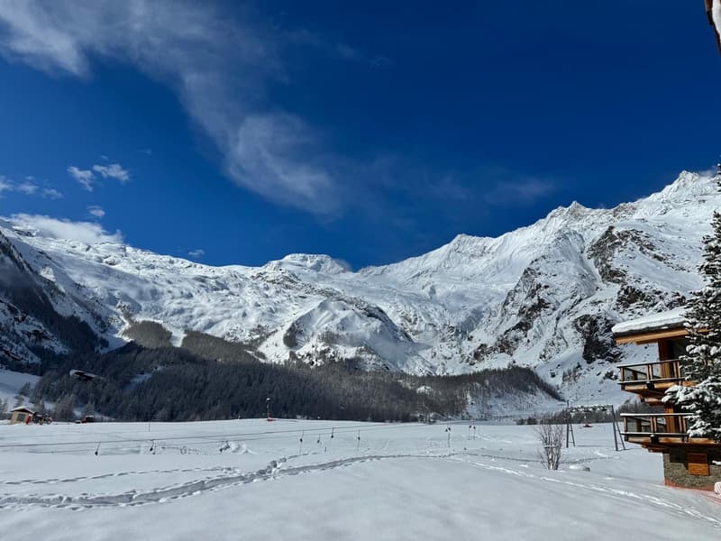 A wooden building with a balcony, surrounded by snowy mountains, with ski lifts visible in the distance