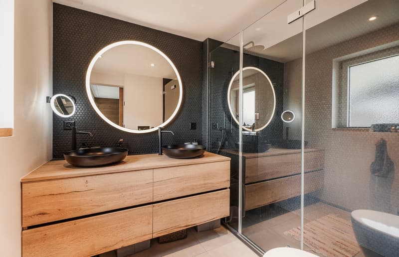 modern bathroom with a wooden vanity with two basins, round mirrors, glass shower enclosure, tiled flooring, white toilet, bathtub, and black hexagonal tiles on the walls
