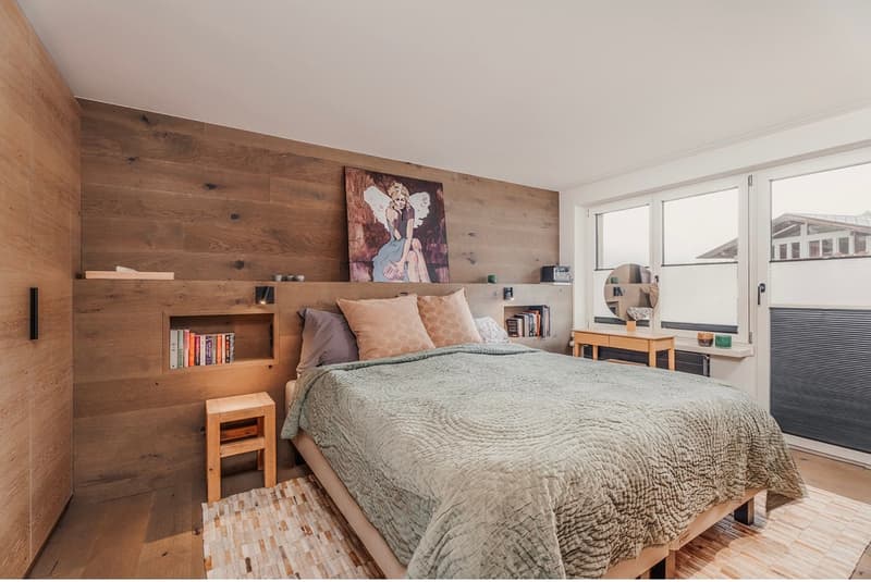 Double bed with wooden frame, headboard wall made of wooden planks, shelves with books, stool, rug, nightstand with mirror, open doors to balcony