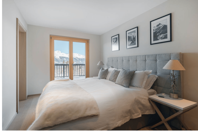 Bedroom with large bed, large windows, balcony, grey upholstered headboard, lamps, white nightstand, white walls, wooden flooring, two framed landscape paintings