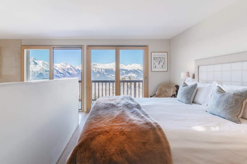 large bed with white sheets, fur blanket, various pillows, sliding glass doors to balcony, picture frame, white walls, tiled floor, modern decor