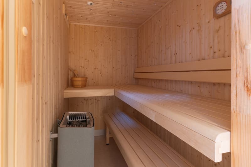 Wooden sauna room with two benches, wood paneled walls, heating element, wooden bowl, clock