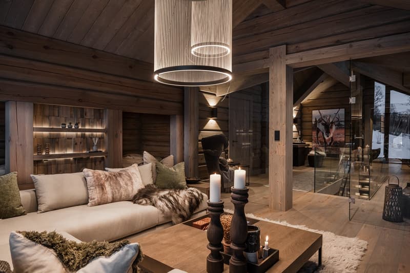 Cozy interior with a wood-paneled wall, fireplace, a beige couch with several fur pillows, center table with candles and a bowl of food, lamps, a wooden chair, and a glass wall with a view to the indoor stair.
