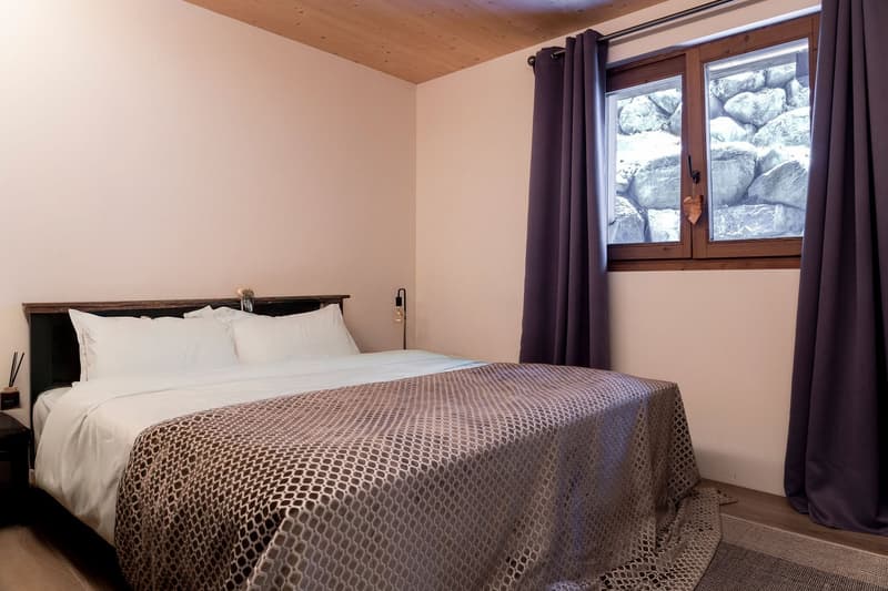 A bedroom with a bed, pillows, and a blanket. A nightstand with a lamp and a heart-shaped decoration is on the left side. A window with curtains is on the right side, and a rug is on the floor.