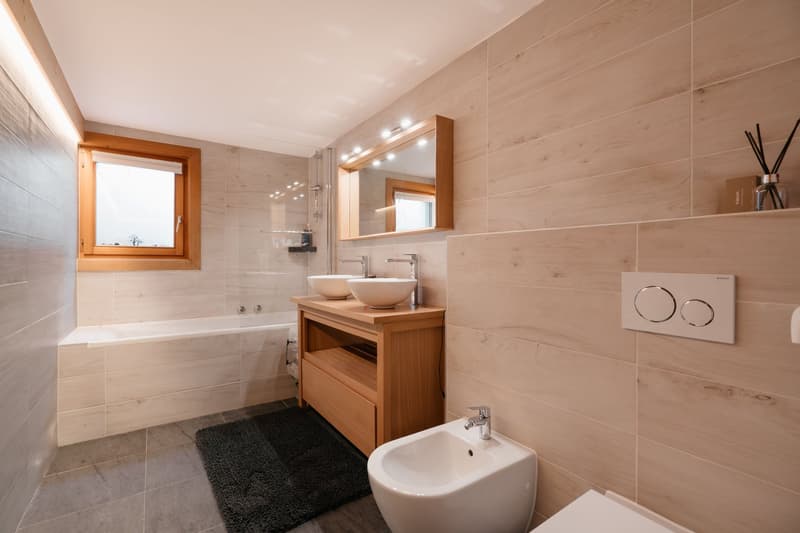 Bathroom with bathtub, double sink, mirror, toilet, bidet, wooden cabinet, tiled walls, window