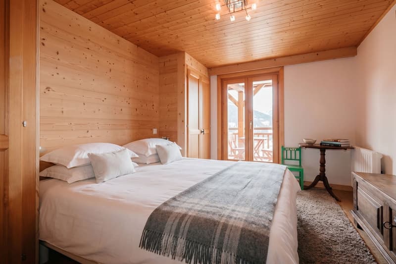 wooden walls, bed with white sheets and pillows, blanket, rug, table with books, green chair, radiator, balcony with chairs