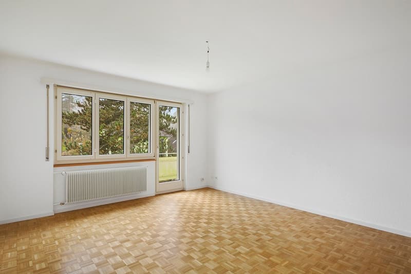 Spacious room with large windows overlooking a garden, hardwood parquet flooring, and a radiator on the wall.